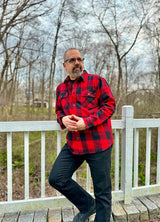 A man leaning against a fence in a red buffalo plaid men's heavyweight flannel plaid shirt